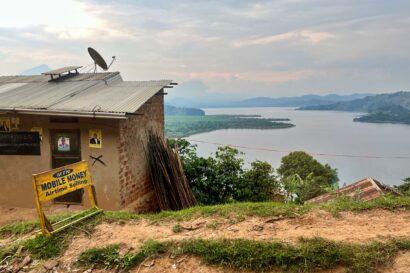 Ugandan landscape shot with a small hut in left foreground, lakes centre and right background and a yellow sign that read mobile money airtime selling in front of the hut