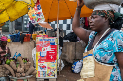 Ghana: A woman carries fruit on her head as she passes a mobile money stand in the Tesano neighborhood.