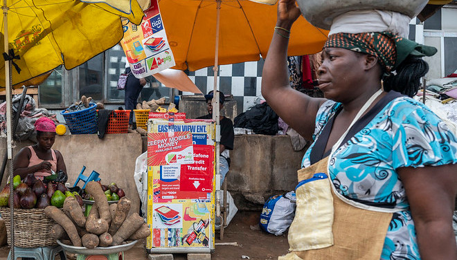 Ghana: A woman carries fruit on her head as she passes a mobile money stand in the Tesano neighborhood.