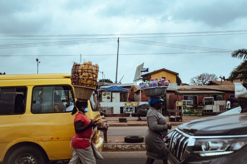 Two market traders walking by cars with baskets of goods over their heads