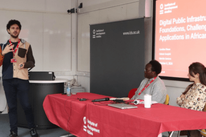 A man is giving a presentation, holding a microphone and standing up at the end of a long table that is covered by a bright red cloth. Behind the table to women are listening to the presentation.