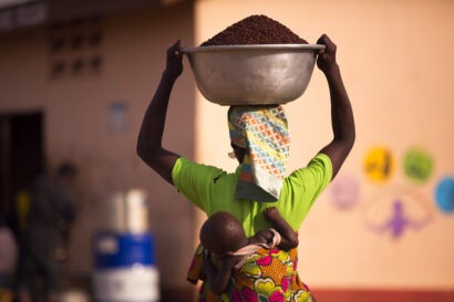 Woman carrying a large bowl of shea nuts with a baby tied behind her back in Ghana.
