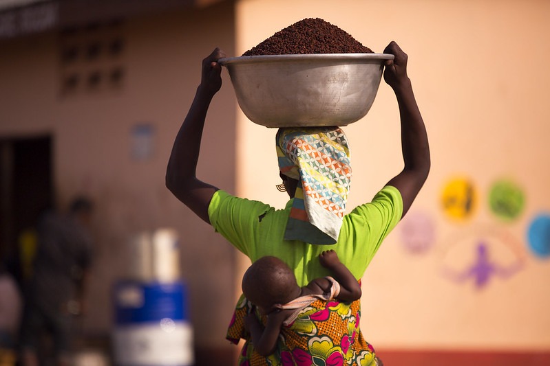 Woman carrying a large bowl of shea nuts with a baby tied behind her back in Ghana.