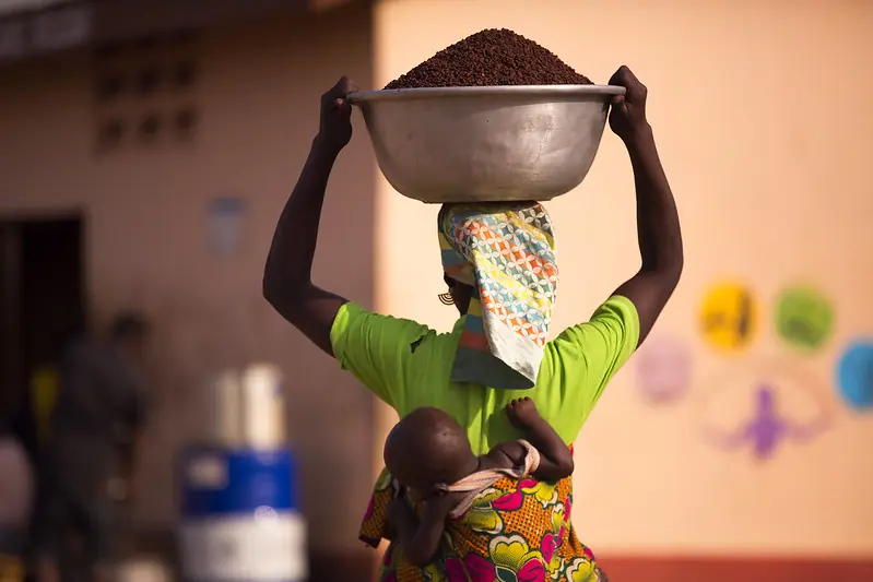 Woman carrying a large bowl of shea nuts with a baby tied behind her back in Ghana.