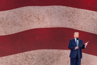 Donald Trump standing behind a microphone with a book in hand and in front of a background of white and red strips.