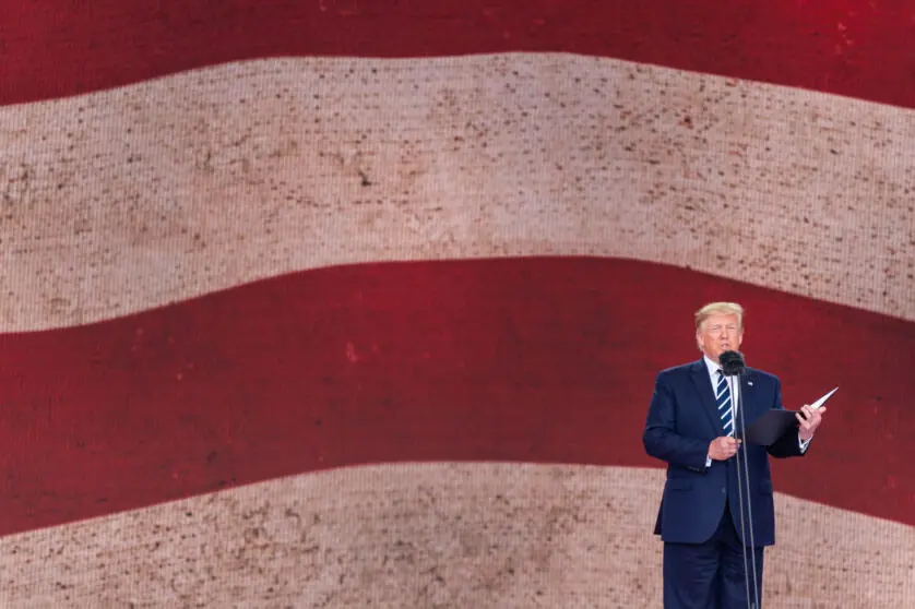 Donald Trump standing behind a microphone with a book in hand and in front of a background of white and red strips.