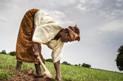 A black woman in modest clothing is bending down towards the ground with a gardening tool in her hand to tend the soil covered in grass.