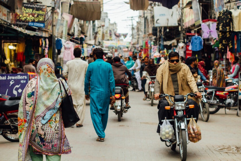 Busy market in Wazirabad, Pakistan, with people walking in the middle road and a man on a motorbike is facing the camera. To the left and right of the main street, market stalls with clothes on display hanging.