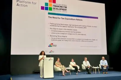 A woman stands behind a lectern, givign a presentation, while a panel of four speakers sit and wait to her right.