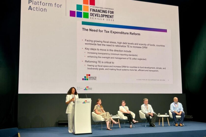A woman stands behind a lectern, givign a presentation, while a panel of four speakers sit and wait to her right.