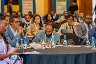 Man in the middle of the frame sitting on a table in a conference room with people around him either in the background or sitting near him on the same table. The man is not looking at the camera but is focused writing on a paper in front of him.
