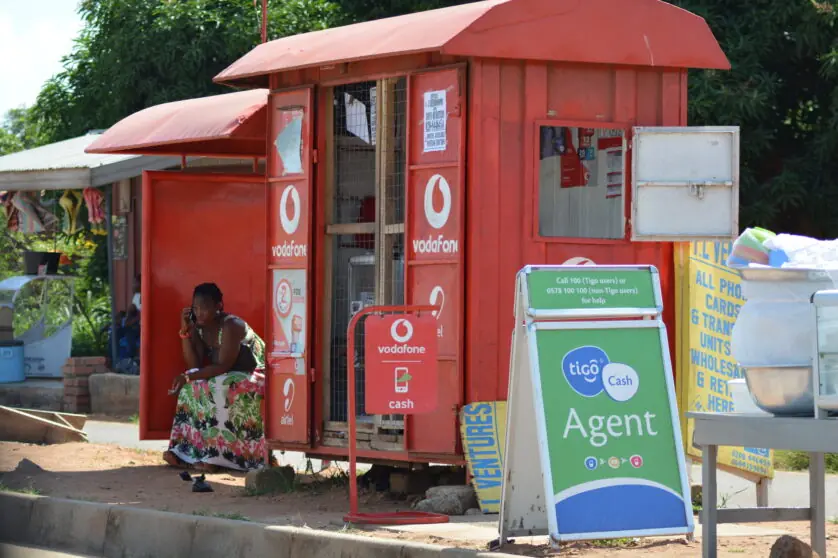 A woman sitting beside a red Vodafone and Tigo Cash mobile money kiosk in Ghana, representing the growing role of digital payment systems in Africa’s digital public infrastructure.