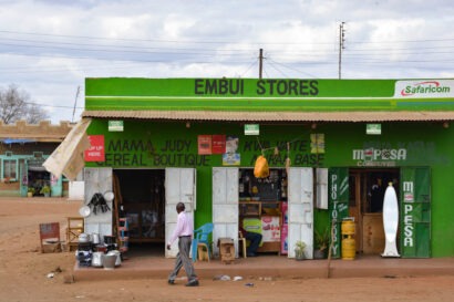 A man walks past a bright green roadside shop in Kenya with Safaricom and M-Pesa signs, illustrating how digital payments support Nigeria digital public infrastructure for taxation across Africa.