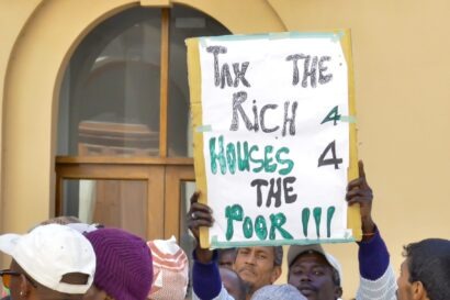 Close up picture of a black man in a street protest in Africa about taxing the wealthy. A sign reads: Tax the rich 4 houses 4 the poor