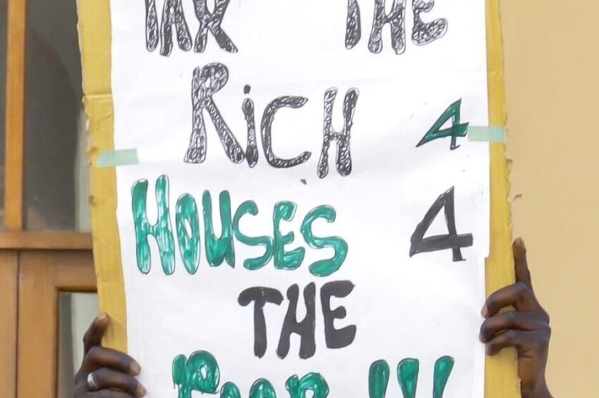 Close up picture of a black man in a street protest in Africa about taxing the wealthy. A sign reads: Tax the rich 4 houses 4 the poor