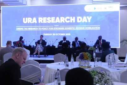 Digital banner displaying the words URA Research Day behind five speakers sitting on a stage facing a crowd of people who are taking notes.