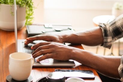 Close up shot on the hands of an African person person on their laptop which is placed on a wooden desk next to a mug.