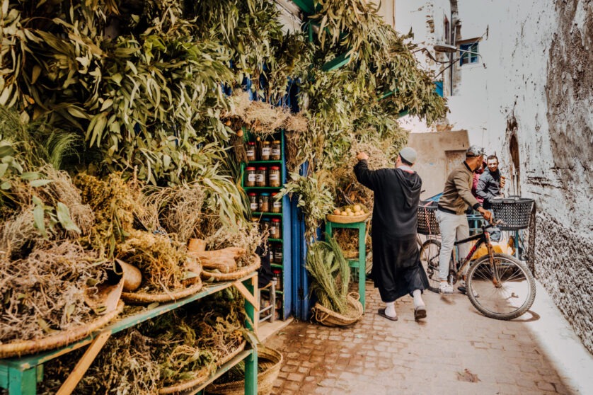 A narrow street market in Morocco with dried herbs and plants displayed on shelves, a man in traditional clothing browsing, and two men with a bicycle nearby.