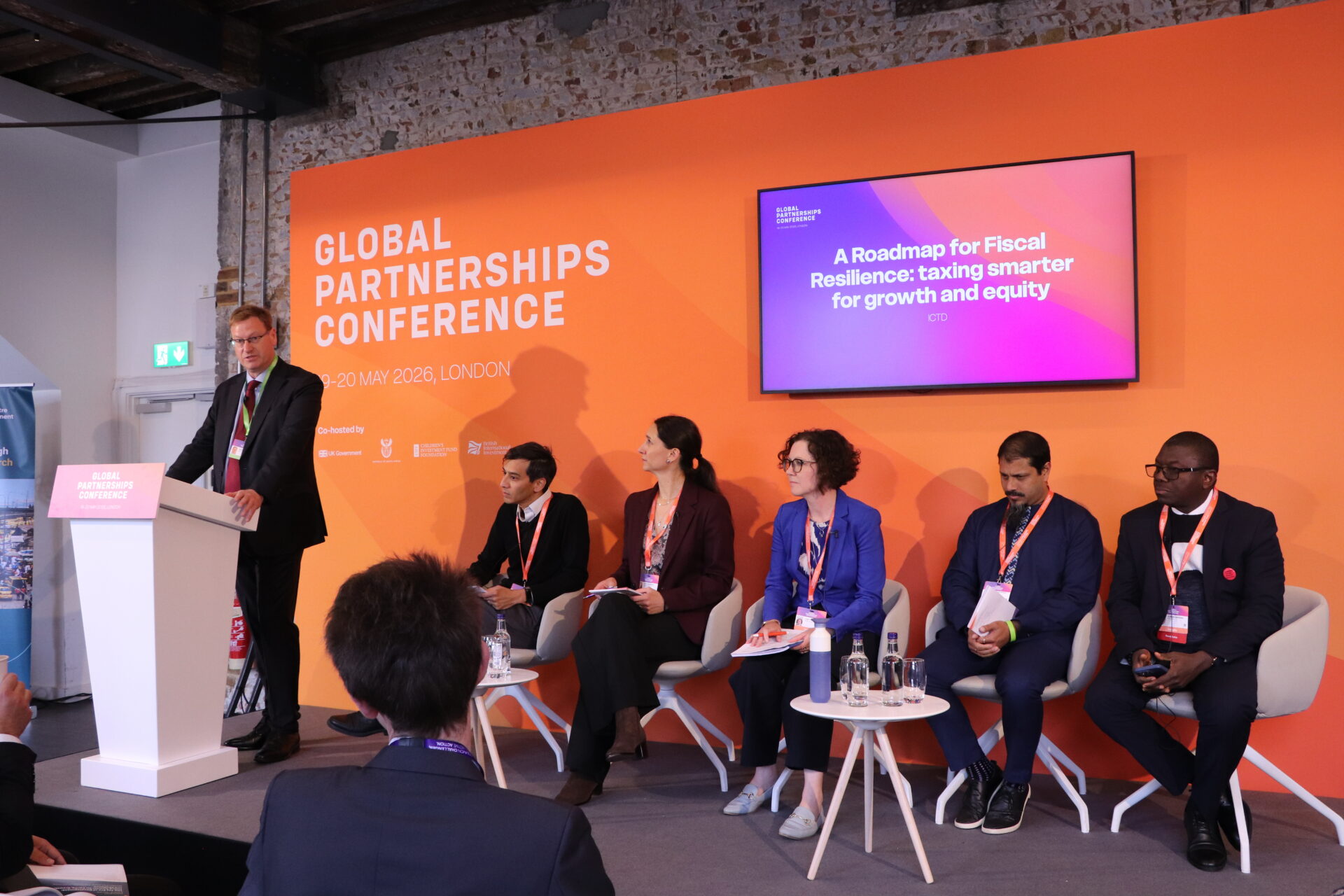 View of the stage at the FCDO Global Partnerships Conference with five seated speakers looking at the Scott Caldwell from FCDO providing the opening remarks