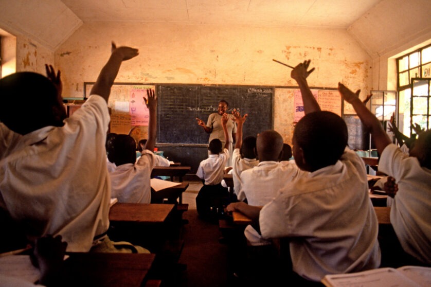 Young students in white uniform raising their hands in the foreground, facing a teacher at the front of the classroom.