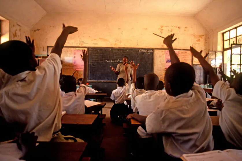 Young students in white uniform raising their hands in the foreground, facing a teacher at the front of the classroom.