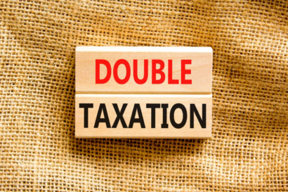 Rectangular wooden block with text 'DOUBLE TAXATION' in red and black on top of a brown fabric.