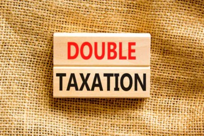 Rectangular wooden block with text 'DOUBLE TAXATION' in red and black on top of a brown fabric.