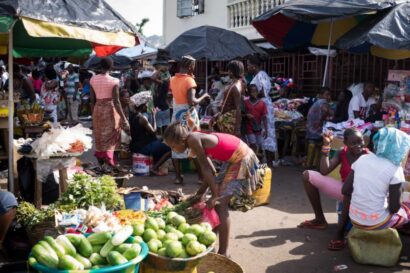 Busy outdoor market scene with vendors selling fresh produce and shoppers browsing under colorful umbrellas.